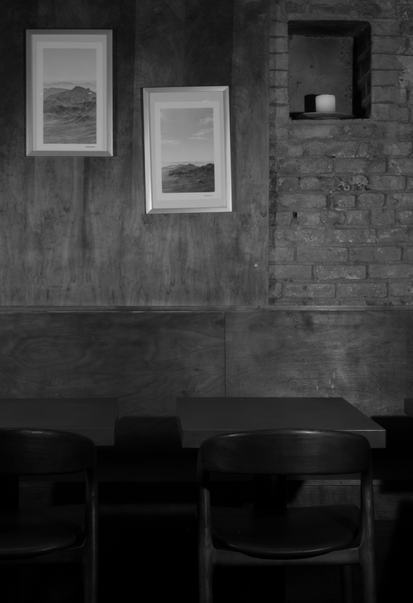 Corima banquette — framed landscape prints on a wood-panel and exposed-brick wall with a candle niche above the tables