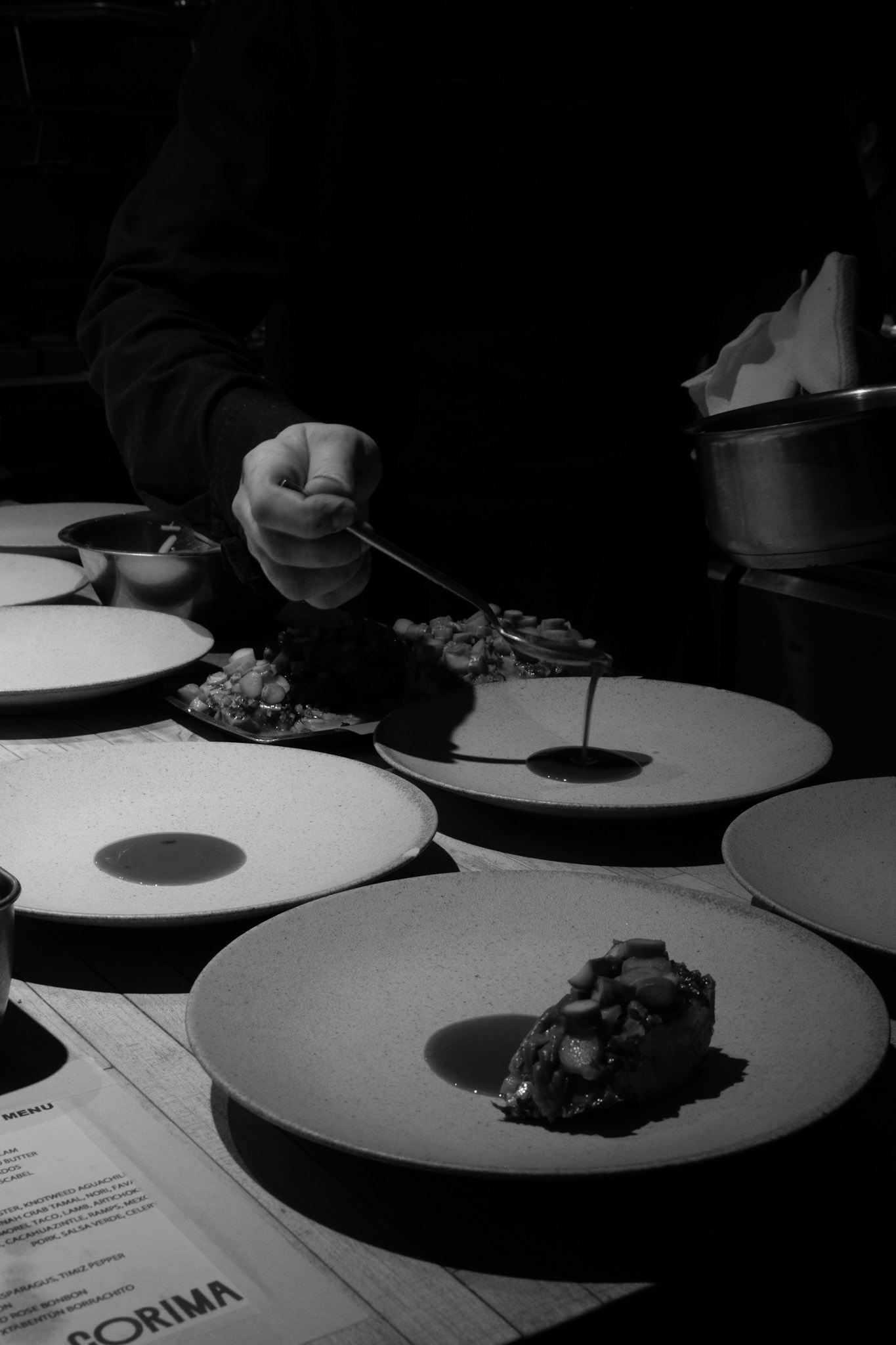A Corima cook spooning sauce and garnish onto speckled ceramic plates at the pass