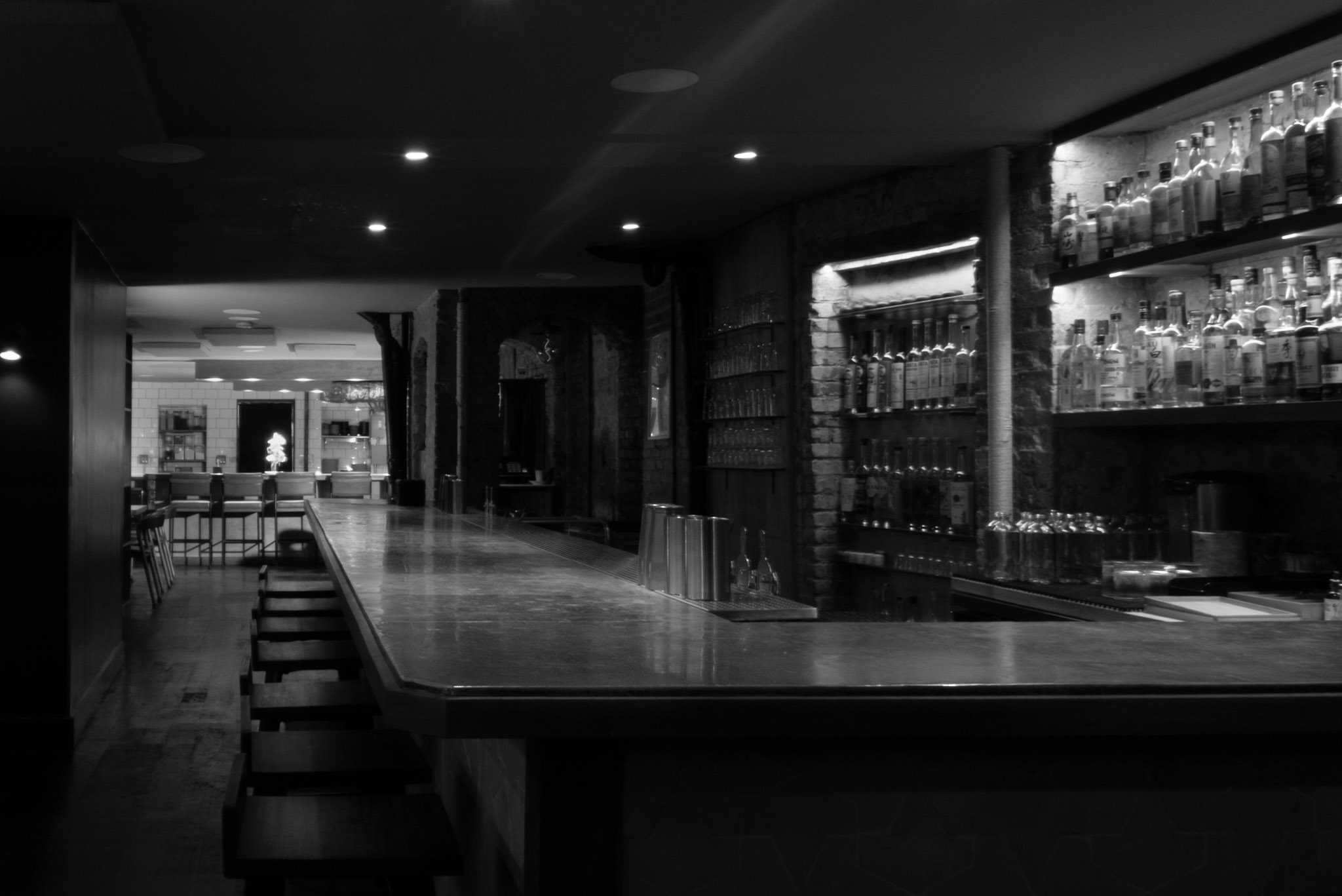 Corima bar — a long counter with stools and backlit shelves of bottles against an exposed brick wall, looking toward the lit dining room
