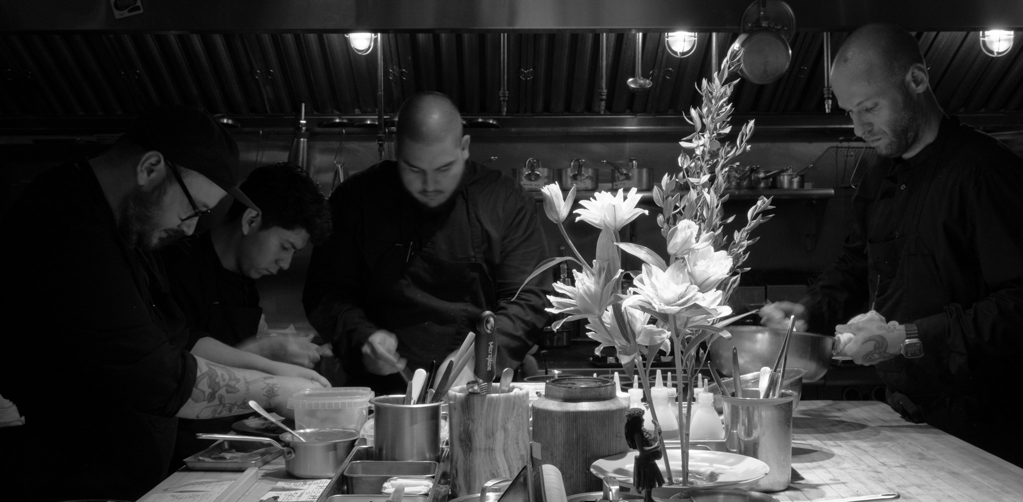 Corima cooks working the line at the pass, with a tall floral arrangement and prep tools in the foreground