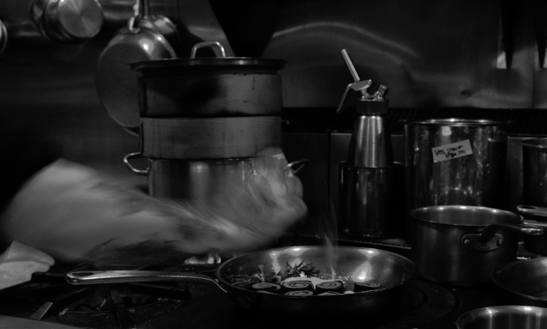 Corima kitchen during service — a chef searing over an open flame beside bamboo steamers and stockpots